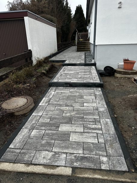 Stilvoll neu angelegter Gehweg mit modernem Betonpflaster und klarer Linienführung Gestalteter Gehweg aus Pflastersteinen, umgeben von Rasen und schmalen Beeten.